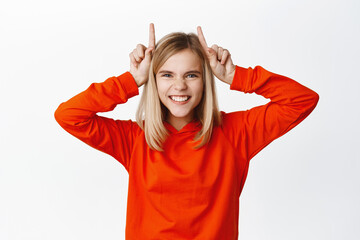 Obraz na płótnie Canvas Happy funny teen girl, kid shows bull horns on head and smiles, fooling around and playing, standing over white background