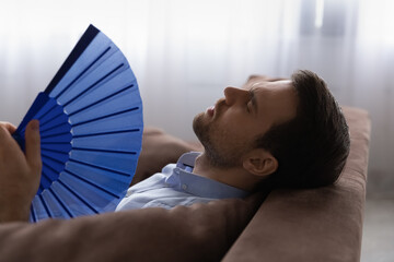 Head shot exhausted man with closed eyes waving paper fan cooling, suffering from overheating o hot...