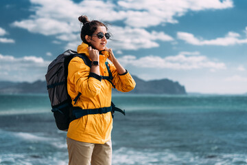A beautiful tourist woman with a backpack on her back standing against the backdrop of a beautiful seascape. Traveler with his tourist backpack is standing against a sea background. Copy space