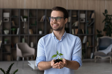 Dreamy smiling man in glasses holding green plant with soil, growing small tree, environment...
