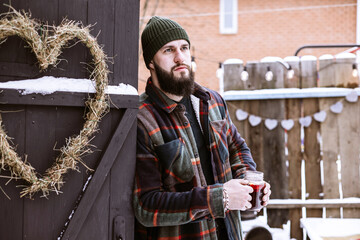 handsome bearded man in stylish winter coat stands outdoor at porch of country house with glass jar of mulled wine, winter spirit concept, decor for valentines day