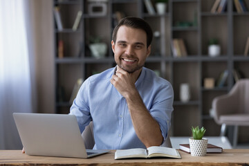 Fototapeta na wymiar Head shot portrait of smiling successful businessman sitting at workplace desk with laptop, happy young man freelancer or student looking at camera, working or studying online, home office