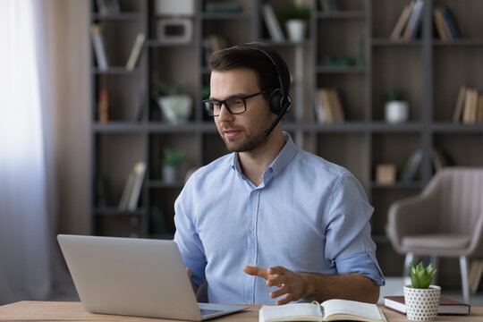 Focused businessman in glasses and wireless headset with microphone making video call, consulting client online, looking at laptop screen, confident man leading internet educational webinar, course