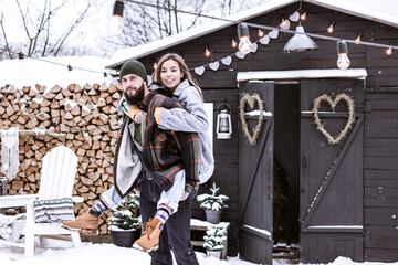 Naklejka premium couple of young woman and man having fun in courtyard of decorated suburban house in winter, concept of Christmas and New Year vacation on farm, family love and support, Valentines Day celebration