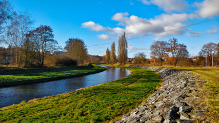 Obraz premium River flows through artificial stream bed made of grass and stony embankment. There are some trees around and the sky has many white clouds.