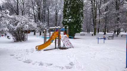Children's slide with inclined plane covered with fresh white snow surrounded by bushes and trees in the public park.