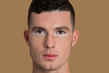 Fototapeta premium Closeup portrait of an attractive young White man with short brown hair, light makeup and white eyeshadow sitting by himself inside a studio with a pecan background wearing a black shirt.