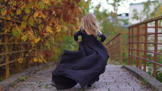 run back Little princess in black dress in autumn. Funny little girl run on street. Spins and laughs. concept of cheerful childhood on street. Running on bridge in halloween. yellow foliage of rowan.