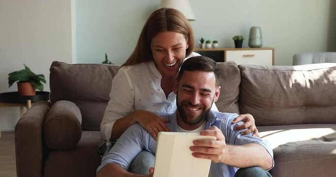 Affectionate girlfriend sit on sofa hug smiling boyfriend from behind look on tablet screen watch funny video with interest laugh on comedy clip. Friendly couple spend weekend at home using pad gadget