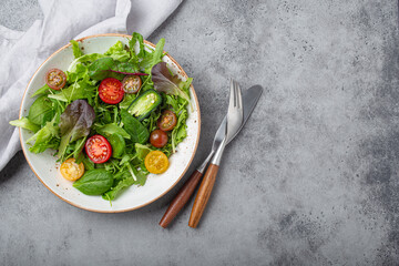 Vegetables healthy salad with red and yellow cherry tomatoes, pepper and green salad leafs on white rustic ceramic plate on gray concrete background top view, healthy food and diet concept, copy space