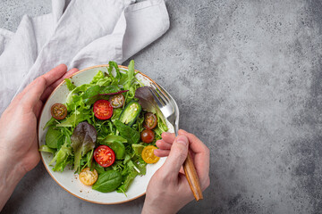 Man eating vegetarian vegetables healthy salad with red and yellow cherry tomatoes and green salad leafs on white plate on gray concrete background top view, healthy food and diet concept, copy space