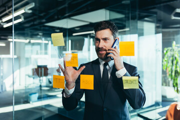 businessman in reflection at a glass task board, a man talking on the phone, hanging plans on...