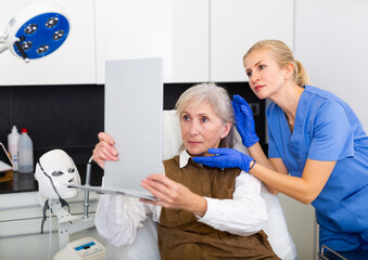 Fototapeta premium Elderly woman looking in mirror during doctor's appointment in cosmetological clinic. Female cosmetologist standing beside.