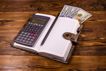 Opened notepad, one hundred dollar banknotes, scientific calculator and pencil on wooden table