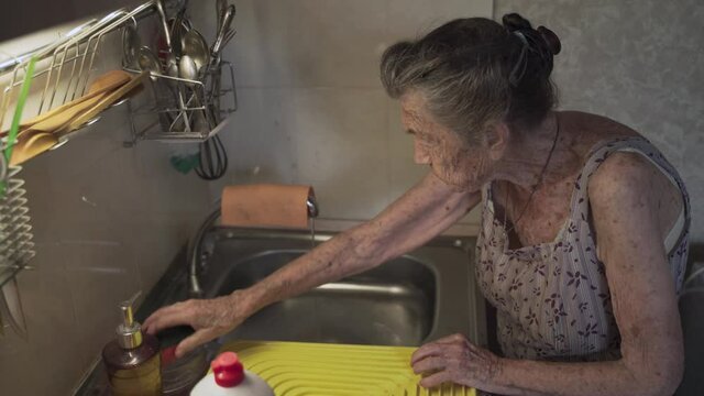 Senior elderly woman washing dishes in kitchen. Sad mature housewife cleans up the old dishes in the kitchen. Upset woman with gray hair 90 years old washing kitchen utensils in a hand wash at home.
