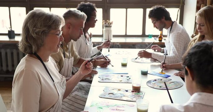 Skilled middle age woman professional drawing teacher give painting master class to diverse talented youth. Happy aged lady fine art school tutor explain art basics to multicultural pupils on workshop