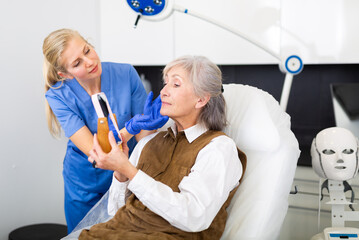 Experienced female cosmetologist giving professional advice before hardware facial skin treatments to aged woman looking at mirror in cosmetological office