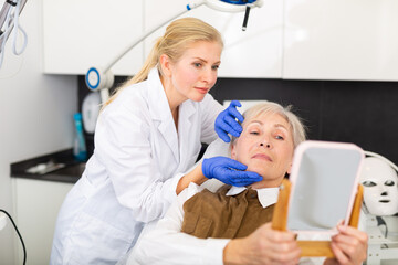 Elderly female patient looking at mirror while professional female cosmetologist explaining her future hardware rejuvenating facial procedure in medical esthetic office