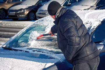 Man scratching ice on car windscreen with ice scraper and brushing snow from car in the morning. Man brushing snow from windscreen of parked car. Cleaning windshield with scraper from ice in winter