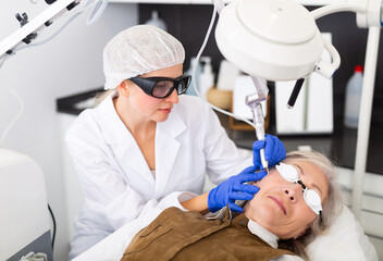 Focused skilled female cosmetologist performing hardware laser treatment to resurfacing and tightening facial skin to senior woman at cosmetology clinic