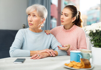 Upset elderly woman sitting at table. Her adult daughter and trying to cheer her up.