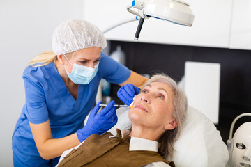 Qualified focused cosmetologist giving beauty injections for face skin rejuvenating and tightening for elderly female client in aesthetic medicine office