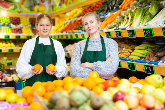Portrait of confident smiling saleswoman standing with trainee salesgirl among shelves with fresh organic fruits and vegetables in grocery store