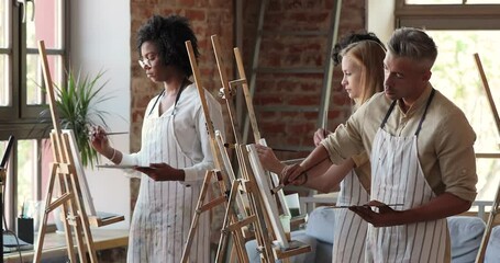 Multiethnic group of men women young artists enjoy working on pictures on canvases using oil acrylic paints. Diverse painters students team hold brushes palettes attend creative workshop at art studio