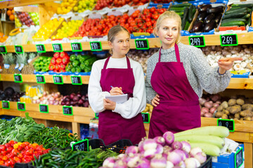 Focused woman grocery store owner giving instructions to young salesgirl, pointing at goods while standing near shelves with fruits and vegetables