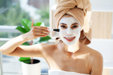 Beautiful young woman with towel wrapped around her head applying face mask at the bathroom