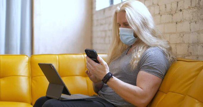 Masked long blonde hair bearded man on yellow vegan leather vinyl couch typing on iPad Pro Magic Keyboard then picking up iPhone 13 Pro Max and texting messaging back - in Cinema 4k (60fps Real Time).