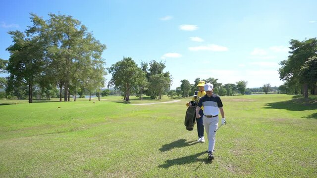 Asian man golfer walking on fairway together with female caddy on the green at golf course in sunny day. Healthy male enjoy outdoor lifestyle activity sport golfing at country club on summer vacation