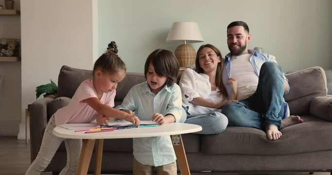 Family of 4 spend cozy evening at home together. Two diverse little children younger daughter elder son preschoolers draw colorful sketches using pencils. Smiling young mom dad cuddle on sofa relax