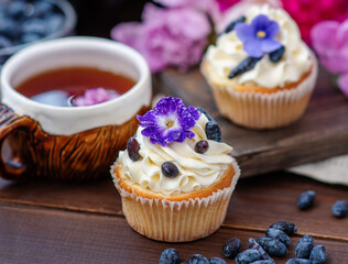 Cupcake with cream decorated with violet flowers lying next to a mug of tea among the flowers of peonies and scattered honeysuckle berries