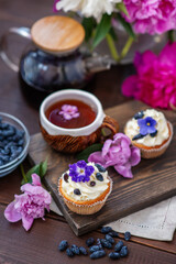 Fototapeta na wymiar Cupcake with cream decorated with violet flowers lying next to a mug of tea among the flowers of peonies and scattered honeysuckle berries
