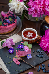 A piece of berry cheesecake lying next to a mug of tea, violet flowers and on a background of peonies and a whole cake