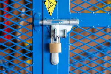 A key padlock is locking on metal fence gate knob of the hand tool storage box (as blurred backgroung). Industrial object photo. Close-up and selective focus. - obrazy, fototapety, plakaty