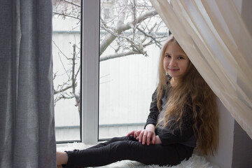 Portrait of a child girl with long hair, sitting on the windowsill.