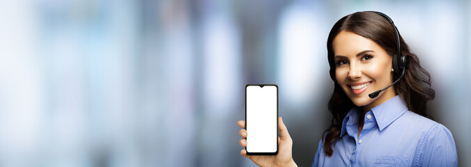 Customer support phone operator in headset holding showing smartphone cell phone mobile with white blank mock up screen blurred office interior background. Consulting and assistance service callcenter