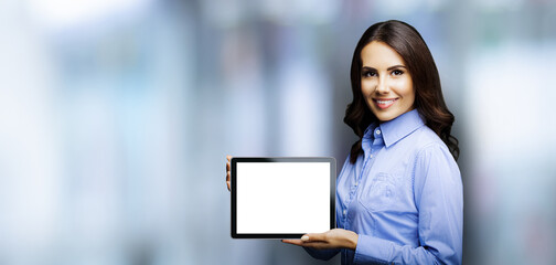 Portrait of cheerfully smiling brunette businesswoman showing mock up blank tablet pc, touchpad, with copy space area, blurred modern office interior background. Confident young business woman indoors