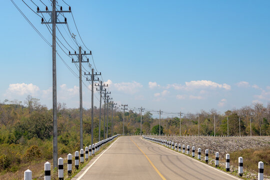 Asphalt road with pebbles stones along the dike under blue sky and white clouds as background, Electric pole and cable line along the street with forest, Traditional countryside in Thailand.