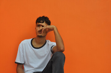 A good looking Indian young guy looking sideways while sitting outside posing against orange wall background with copy space 