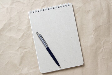 Gray notepad with white coiled spring and pen on a background of beige crumpled craft paper. With empty space for text and design