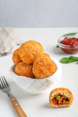 Homemade Arancini Sicilian rice balls or croquettes deep fried and stuffed with minced beef meat in tomato sauce and green peas served in bowl on white wooden table with fork and ketchup. Vertical