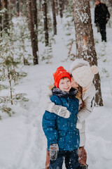 Fototapeta na wymiar mom kisses her son in the winter forest. Mom walks with her son in a snowfall in the forest. Happy winter holidays with snow. Winter walks in the forest. Mom's love for her son.