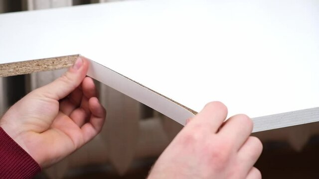 A man glues white melamine edging to the raw edge of wood chipboard furniture.