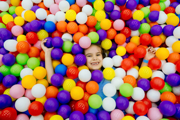 Fototapeta na wymiar Girl kid playing and having a good time in a ball room, little smiling girl playing lying in colorful balls park playground. Happy little girl having fun jumping into the ball pit with colorful balls.