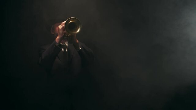 Senior musician in hat and classic suit plays musical trumpet and presses condensate output key in studio on black background with smoke. Concept of jazz, professional instrumental music