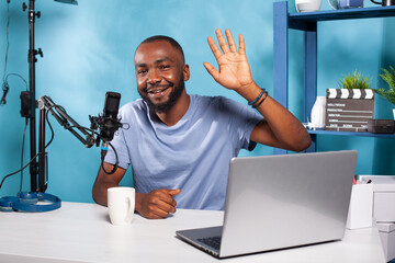 Vlogger saying hello to followers in online morning show sitting at desk with professional microphone and broadcasting setup. Content creator waving hand at audience in front of laptop computer.