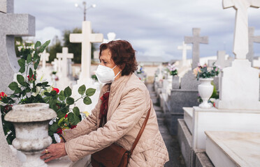 Senior woman wearing protective face mask putting flowers on grave in cemetery